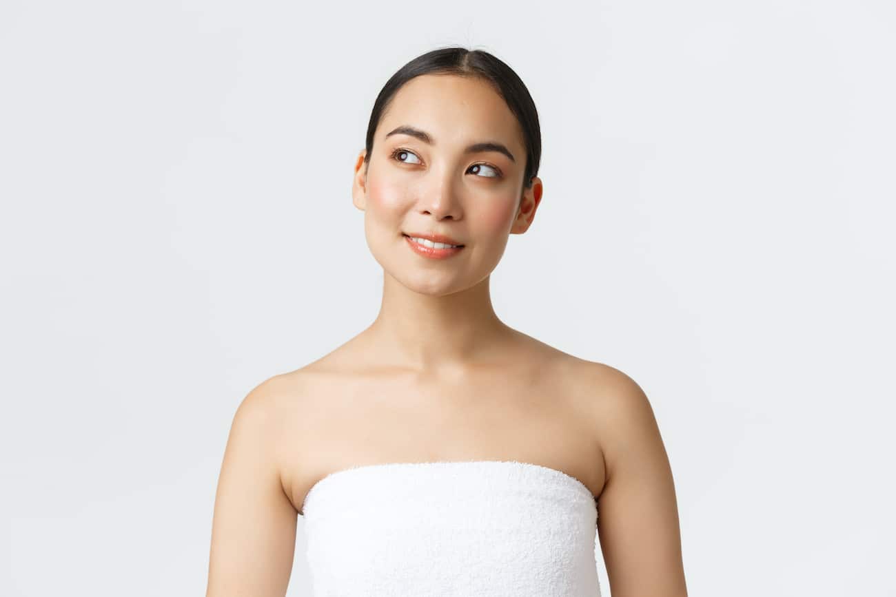 Thoughtful beautiful asian woman with perfect clean skin standing in towel and looking curious upper left corner, thinking, see skincare or haircare product advertisement, visit spa salon